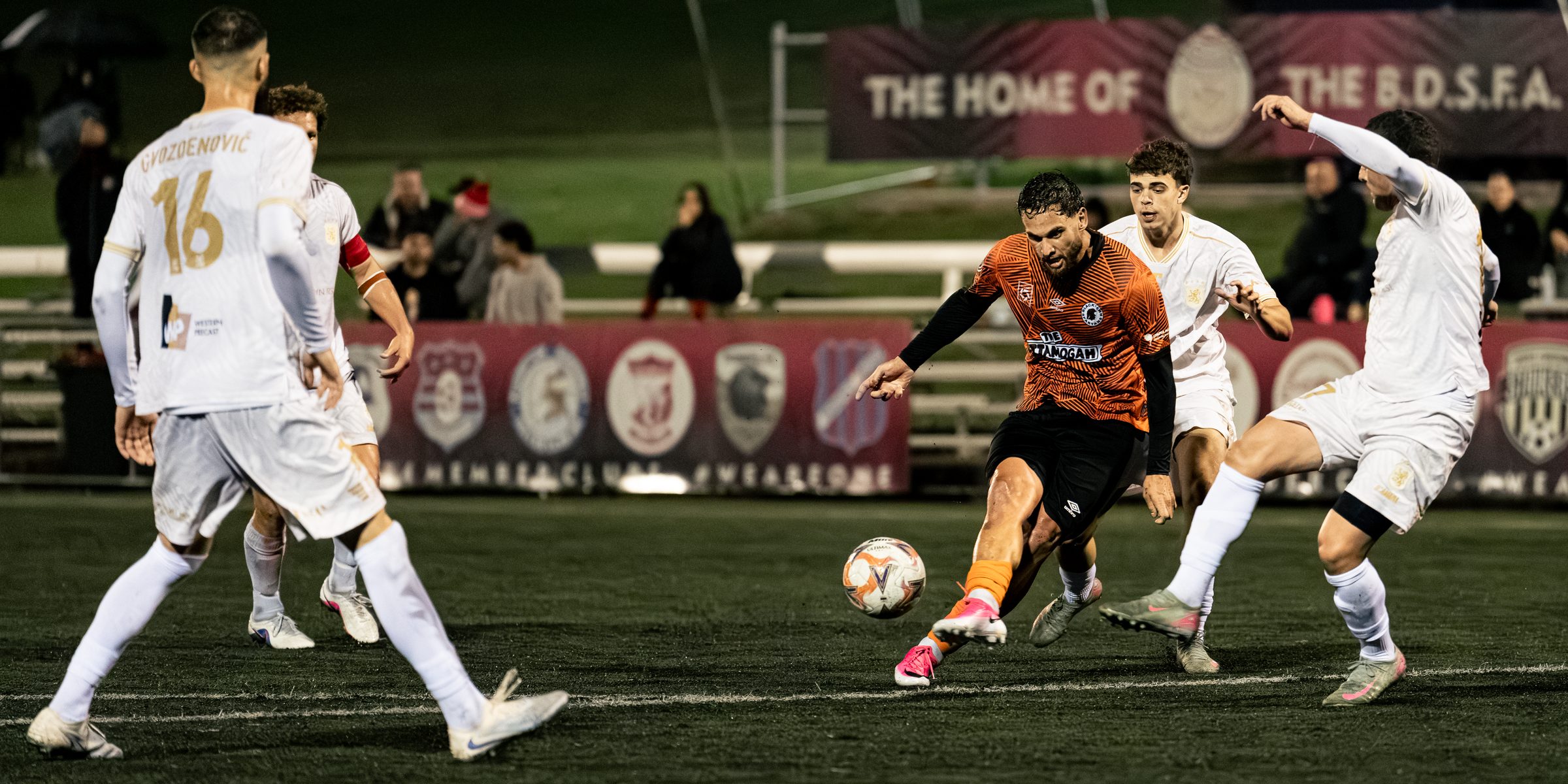 Blacktown Spartans player in orange striking the ball under lights at Blacktown Football Park